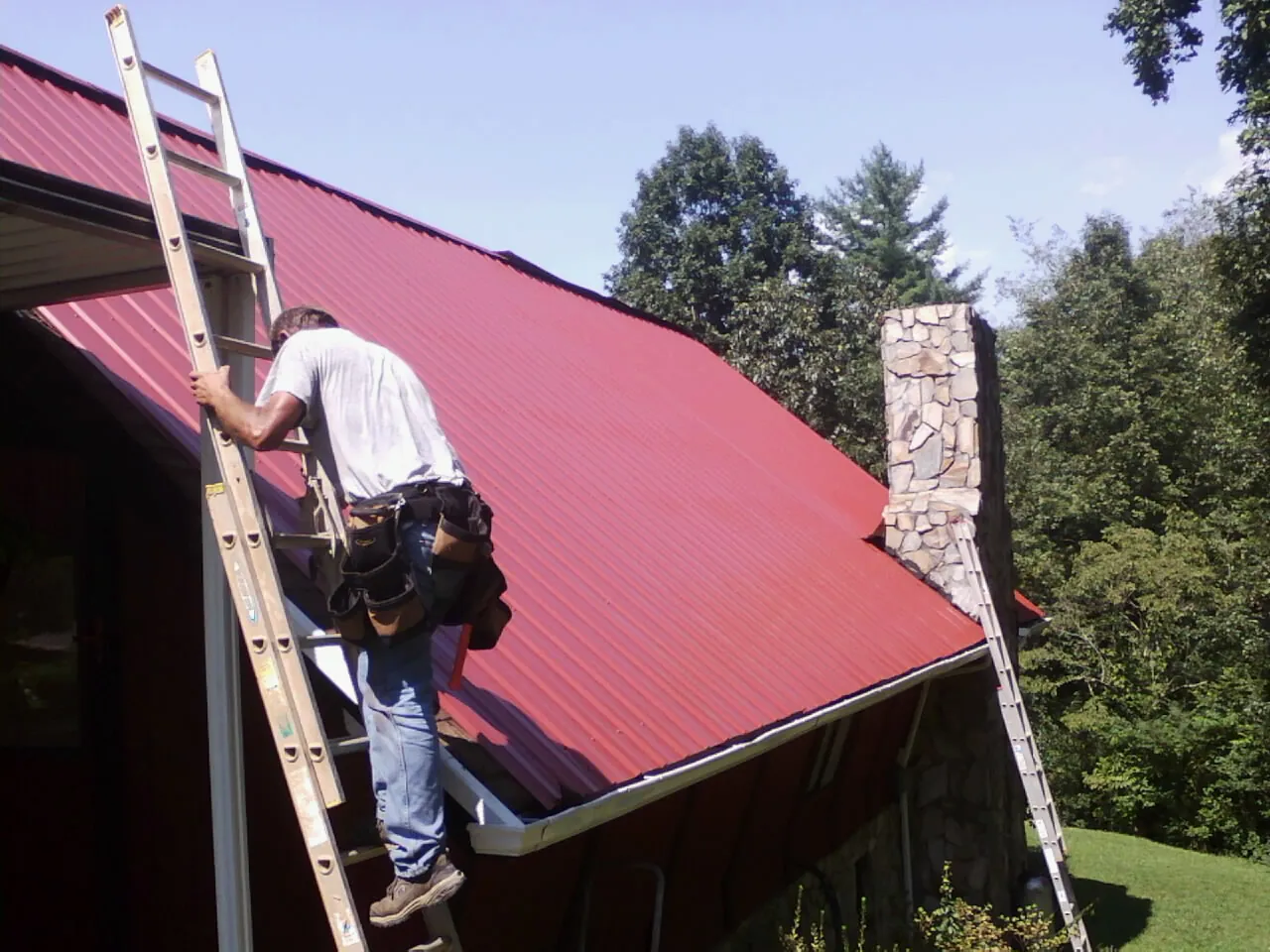 Roofer on a ladder beside a red metal roof and stone chimney — Soffit & Fascia Repair by Roofing Brothers in Monrovia